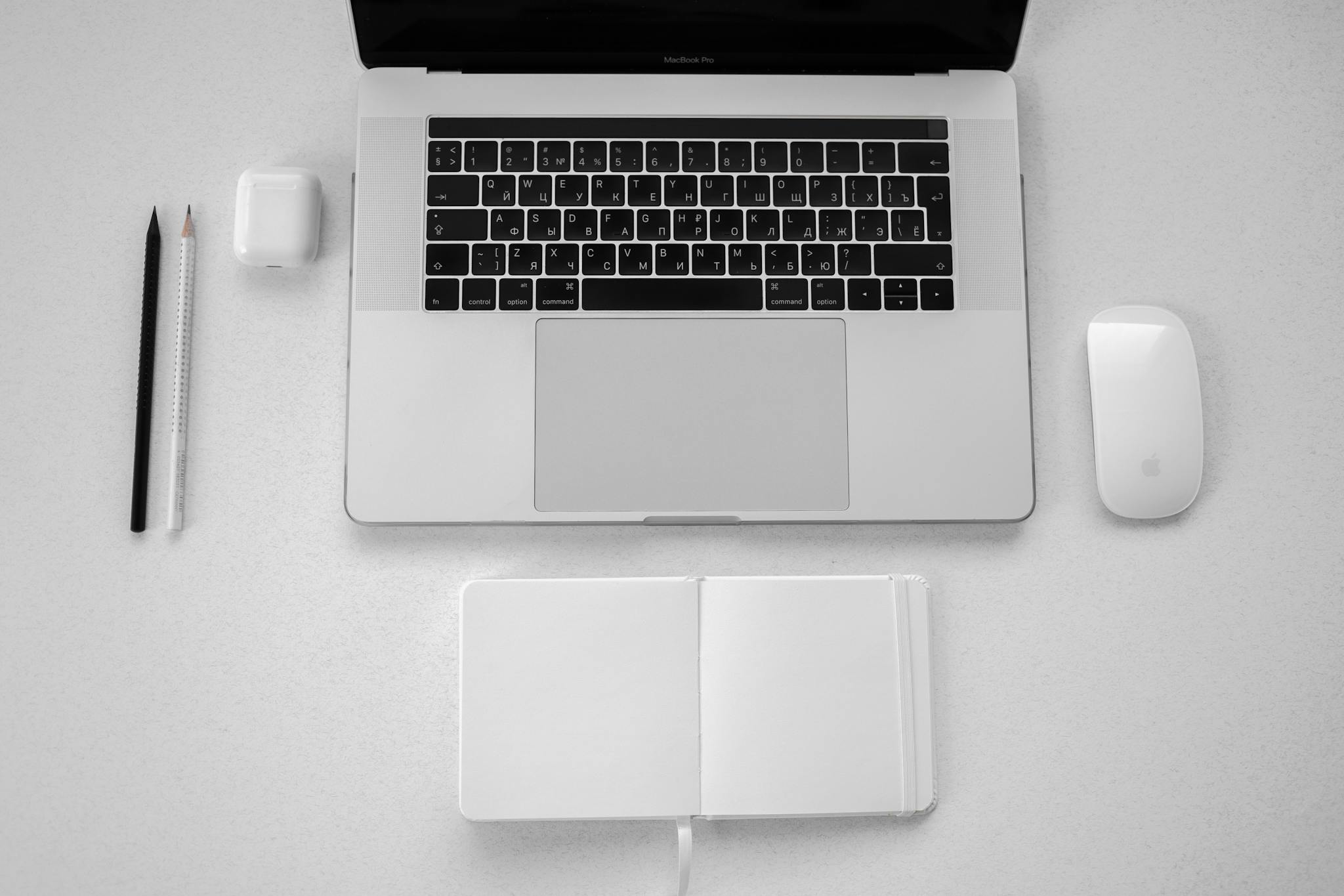 A minimalist top-down view of a workspace featuring a laptop, notebook, pencils, and wireless mouse.