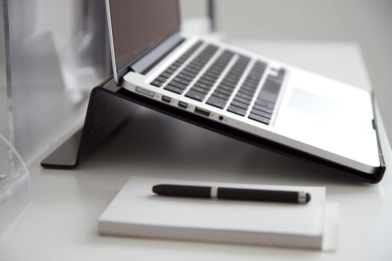 Side view of a laptop on a stand with a notebook and pen on a white desk.