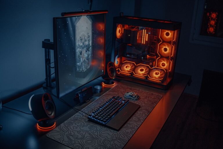 PC de bureau à boîtier vitré éclairé en orange, écran, clavier et enceintes sur un bureau